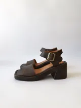 Sandalias de cuero color chocolate, con tira ancha sobre los dedos, pulsera al tobillo con hebilla metálica y taco cuadrado de 6 cm.
