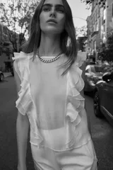 Blusa blanca semitransparente de cuello redondo y sin mangas, con volantes en los hombros y en el frente.