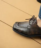Zapato acordonado de cuero vacuno color marrón oscuro, con suela de goma, horma cuadrada y tacón bajo.