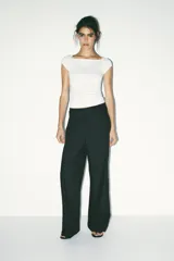 Pantalón negro con finas rayas verticales blancas, de tiro medio y corte ancho. Presenta bolsillos delanteros y traseros, cintura doble tipo bóxer con botones y cierre frontal con cremallera y ganchos metálicos. Detalle de franja marrón en los laterales.