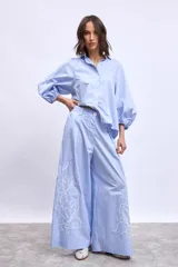 Pantalón ancho de algodón color celeste claro, con bordados de flores blancas en los bajos. Presenta un corte de tiro alto con pliegues frontales y cierre de botón visible.