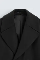 Abrigo negro de corte relajado confeccionado en tejido con 19% de lana. Tiene cuello con solapas de pico, manga larga, bolsillos de vivo en cadera, bolsillo interior, cinturón del mismo tejido en cintura, bajo con abertura en el centro de la espalda y cierre frontal de botonadura cruzada.