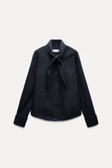 Camisa de denim azul oscuro con cuello con lazos, mangas largas con puños abotonados, bajo redondeado y cierre frontal con botones ocultos.