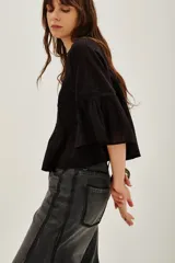 Camisa blanca de algodón con cuello alto y volados, mangas largas abullonadas con puños elásticos. Presenta detalles de encaje y frunces en el frente y la espalda.