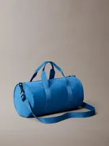 Cartera tipo duffle deportiva de color azul brillante, confeccionada en poliéster. Presenta asas de mano y una correa ajustable para el hombro. En el centro tiene estampado el logo de Calvin Klein en blanco, incluyendo la dirección "West 39th Street New York City". Posee un bolsillo exterior con cierre en el frente.