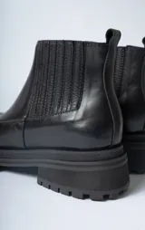 Bota corta tipo Chelsea de cuero negro, con puntera cuadrada y suela track gruesa y dentada. Presenta paneles elásticos acanalados a los lados para facilitar el calce.