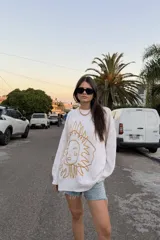 Buzo de tejido de punto color crudo, con cuello redondo y manga larga. Presenta un bordado frontal grande con un diseño abstracto que simula un rostro con rayos solares.