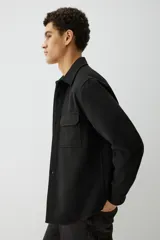 Sobrecamisa negra de corte recto, con cuello clásico, cierre frontal con botones, dos bolsillos de parche con solapa en el pecho, mangas largas con puños abotonados y bajo redondeado.