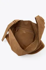 Bolso tipo saca asimétrica color marrón, confeccionado en serraje. Tiene asa bandolera ajustable y cierre con imán. Incluye una cartera con bolsillo extraíble.