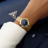 Reloj Guess modelo Clara con caja redonda dorada y bisel decorado con cristales. Esfera azul marino con marcadores de hora en forma de cristal y manecillas doradas. Brazalete de eslabones dorados.
