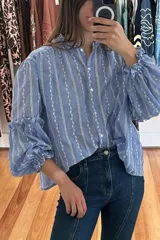 Camisa celeste con rayas verticales blancas y bordados de flores blancas. Tiene cuello mao, mangas largas abullonadas con volados en los puños y cierre frontal con botones de nácar.