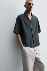 Camisa de denim de algodón, color beige claro, con corte relajado, cuello bowling y manga corta. Presenta cierre frontal con botones a presión y un pequeño bordado a tono en el pecho.