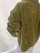 Buzo de pana fina color verde oliva, con diseño de corte holgado y mangas largas. Presenta una textura de cordón vertical característica de la pana y un pequeño parche decorativo en la parte superior de la espalda.
