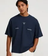 Remera de algodón azul marino, corte oversized, con cuello redondo y mangas cortas. Presenta un pequeño estampado en el pecho con texto y un logo circular, y un estampado más grande con el texto "Broad Plans" en la parte inferior derecha del frente.