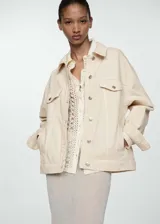 Chaqueta color crema estilo tejana vaquera, de corte oversize, con cuello camisero, cierre frontal de botones, mangas largas con puños abotonados, dos bolsillos de parche con solapa en el pecho y bolsillos laterales.