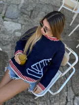 Sweater azul marino tejido a mano en lana merino 100% natural, con cuello, puños y ruedo acanalados en blanco y rojo. Presenta un diseño de barco a vela en blanco en el frente.