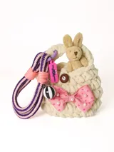 Cartera tejida color crema con asa de soga a rayas violetas y rosadas. Incluye un conejo de peluche beige en su interior y está decorada con un moño rosa con lunares blancos y un botón marrón.