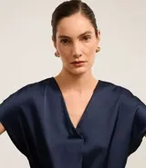 Blusa color café confeccionada en poliéster y elastano, con acabado satinado, escote V y mangas cortas. Presenta pliegues delanteros.