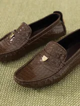 Mocasín de cuero vacuno color marrón con textura croco y acabado brillante. Incluye aplique metálico en forma de corazón sobre el frente y suela de goma.