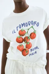 Remera blanca de corte holgado con estampado de tomates y texto en azul. Tiene cuello redondo ribeteado acanalado y hombros caídos.