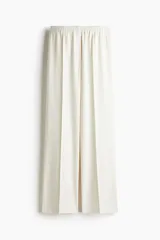 Pantalón de sarga color crudo con finas rayas verticales en gris, de tiro alto con cintura elástica y cierre decorativo. Tiene bolsillos laterales discretos y piernas anchas con pliegues cosidos adelante y pliegues planchados atrás.