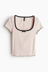 Top de punto acanalado color rosa claro, de manga corta y corte ajustado. Tiene escote cuadrado con ribete negro, moño de satén negro en el centro del busto y ribetes sobrehilados negros en puños y bajo.