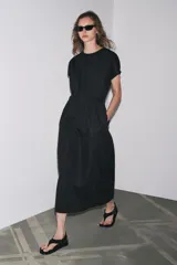 Vestido midi color marrón confeccionado en hilatura con mezcla de algodón y lino. Tiene cuello redondo, manga corta caída y detalle de pliegues en la cintura. Cierre en la espalda con abertura y botón.