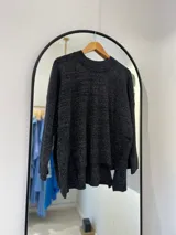 Sweater negro de punto con hilado metalizado, cuello a la base y manga pegada. Delantera más corta que la parte trasera, con abertura en los laterales.