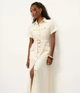 Vestido midi color crema, tipo camisero, con cuello, manga corta y cierre frontal de botones. Presenta bolsillos utilitarios en el pecho y la cadera, y un cinturón ajustable con hebilla redonda de madera. Confeccionado en sarga de algodón.