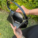 Cartera de hombro negra, estilo baguette, con acabado charol y textura de cocodrilo. Posee correa corta ajustable con detalle de nudo y herrajes dorados.