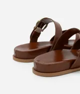 Sandalia plana marrón de cuero sintético, con punta redonda, tiras cruzadas y hebilla dorada lateral.