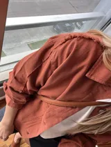 Sobre camisa color ladrillo con cuello camisero, cierre frontal con botones metálicos y bolsillos de parche en el pecho.