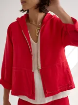 Chaqueta roja de lino, corte holgado, con capucha, cierre frontal con cremallera dorada y bolsillos laterales.