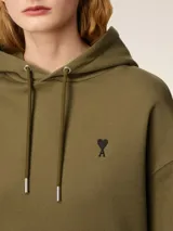 Buzo con capucha color verde oliva, con cordones ajustables y bolsillo tipo canguro. Logo bordado de corazón negro en el pecho. Corte holgado.