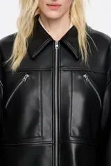Chaqueta negra de cuero sintético con cuello camisero, cierre frontal con cremallera, bolsillos con cremallera en el pecho y mangas largas con cremalleras en los puños.