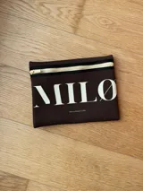 Clutch de neopreno color marrón oscuro con cierre de cremallera metálica dorada. Presenta la inscripción "MILO" en letras blancas de gran tamaño en el frente.