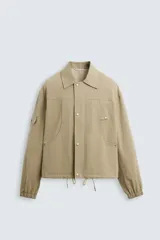 Cazadora color arena de corte relaxed fit, confeccionada en tejido de algodón. Cuenta con cuello solapa, manga larga con bolsillo multifunción, bolsillos frontales de plastrón, bolsillo interior, acabados con elástico y bajo ajustable con cordón. Cierre frontal con cremallera oculta por solapa con botones a presión.