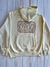 Hoodie color crema con capucha y cordón ajustable. Estampado en la espalda con un paisaje de montañas, árboles y un oso.