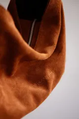 Bolso de hombro tipo hobo de cuero gamuzado color camel, con correa ancha ajustable con hebilla metálica plateada. La correa presenta un estampado de vaca (cow print) en tonos blanco y marrón.