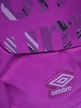 Mochila de color morado intenso con estampado grande y abstracto de la palabra "Umbro" en tonos de morado más claro y negro. Presenta un bolsillo frontal con cierre y un bolsillo lateral de malla para botella. Incluye asa superior y logo Umbro bordado en la parte inferior frontal.