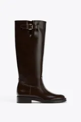 Bota alta de cuero color marrón oscuro, estilo hípica, con hebilla dorada decorativa en la parte superior de la caña.