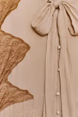 Vestido midi color beige con detalles de encaje marrón, cuello alto con lazo, escote pico, manga larga y botones frontales.
