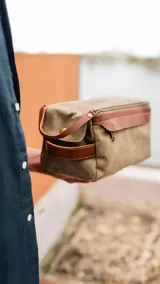 Neceser verde de tela canvas con detalles en cuero marrón. Tiene doble cierre superior, bolsillos internos con cierre y abiertos, bolsillo frontal con solapa y asa lateral.