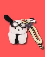 Llavero con forma de perro de peluche blanco con orejas negras, gafas de aviador y corbata negra. Incluye una cuerda trenzada beige y azul con detalles en rosa y verde.