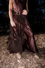 Bolso tote de gran tamaño, color marrón oscuro, confeccionado en material gamuzado. Presenta tiras de hombro largas con flecos colgantes, detalles de hebillas metálicas en los laterales y un parche de cuero marrón claro cosido en el frente.