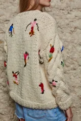 Sweater de lana color crema, tejido a mano con cuello redondo y mangas largas con trenzas. Presenta bordados de figuras esquiando en varios colores.
