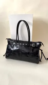 Bolso grande de cuero marrón con textura tipo cocodrilo, estructura rígida y asas para llevar al hombro. Tiene una tira con tachas y hebillas en cada extremo, también lleva lazos en los costados.