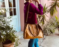 Bolso de mano grande de cuero color marrón, con doble asa corta y correa larga desmontable. Tiene cierre superior y detalles de flecos en el frente.