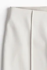 Pantalón tobillero color crudo de sarga con tiro alto, elástico oculto en la cintura y cierre lateral oculto. Piernas ajustadas con bajo levemente acampanado y pliegues frontales.