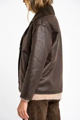 Chaqueta tipo gamulán de calce oversize en color marrón oscuro, con exterior de poliéster y forro de corderito sintético. Presenta cuello de solapa ancha y cierre frontal con botones.